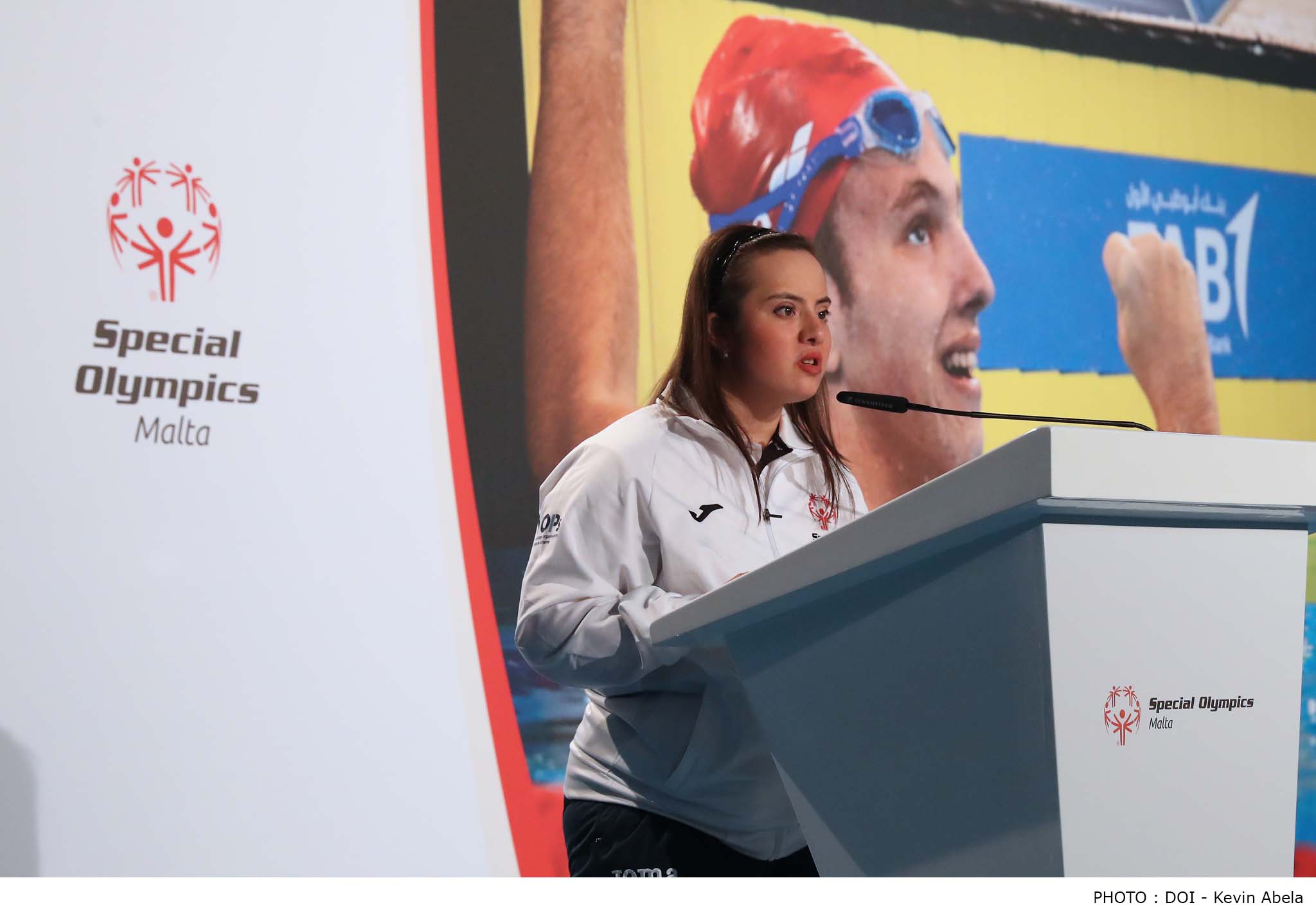 : A young woman speaks into a microphone on a podium with the Special Olympics Malta logo on the podium and in the background. 