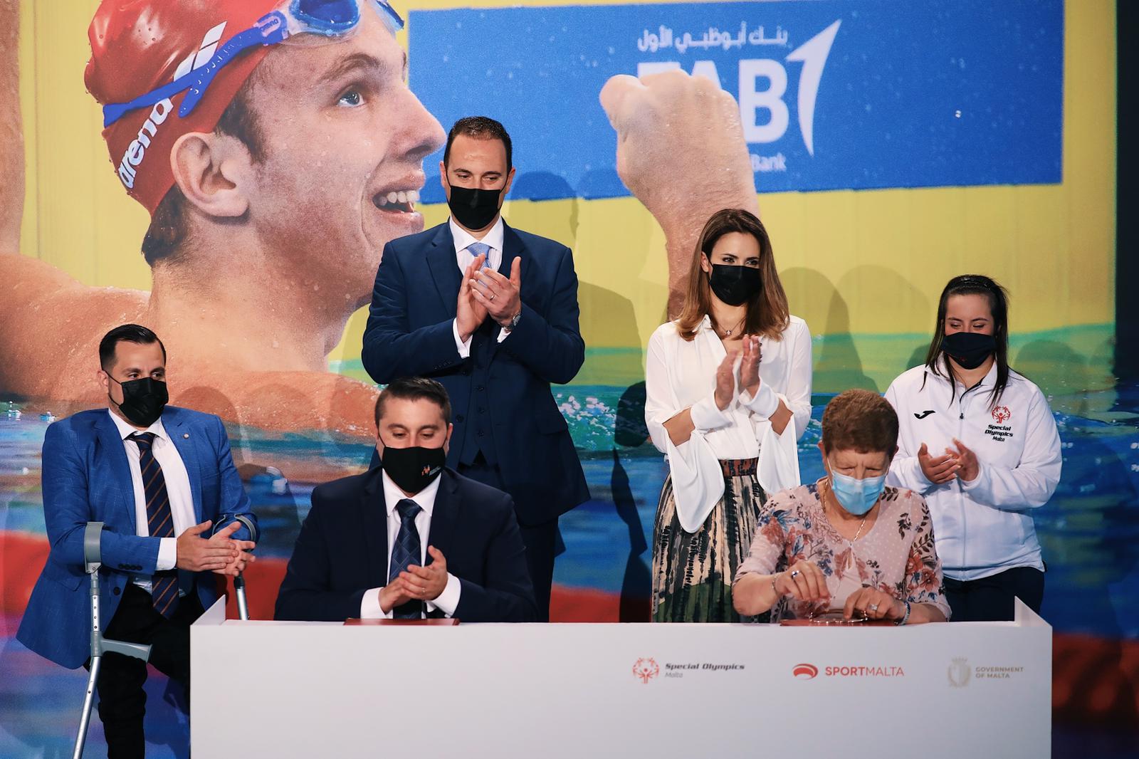 Two people sit behind a desk looking at a document in with a group of four people behind them clapping. Everyone is wearing a face mask. 